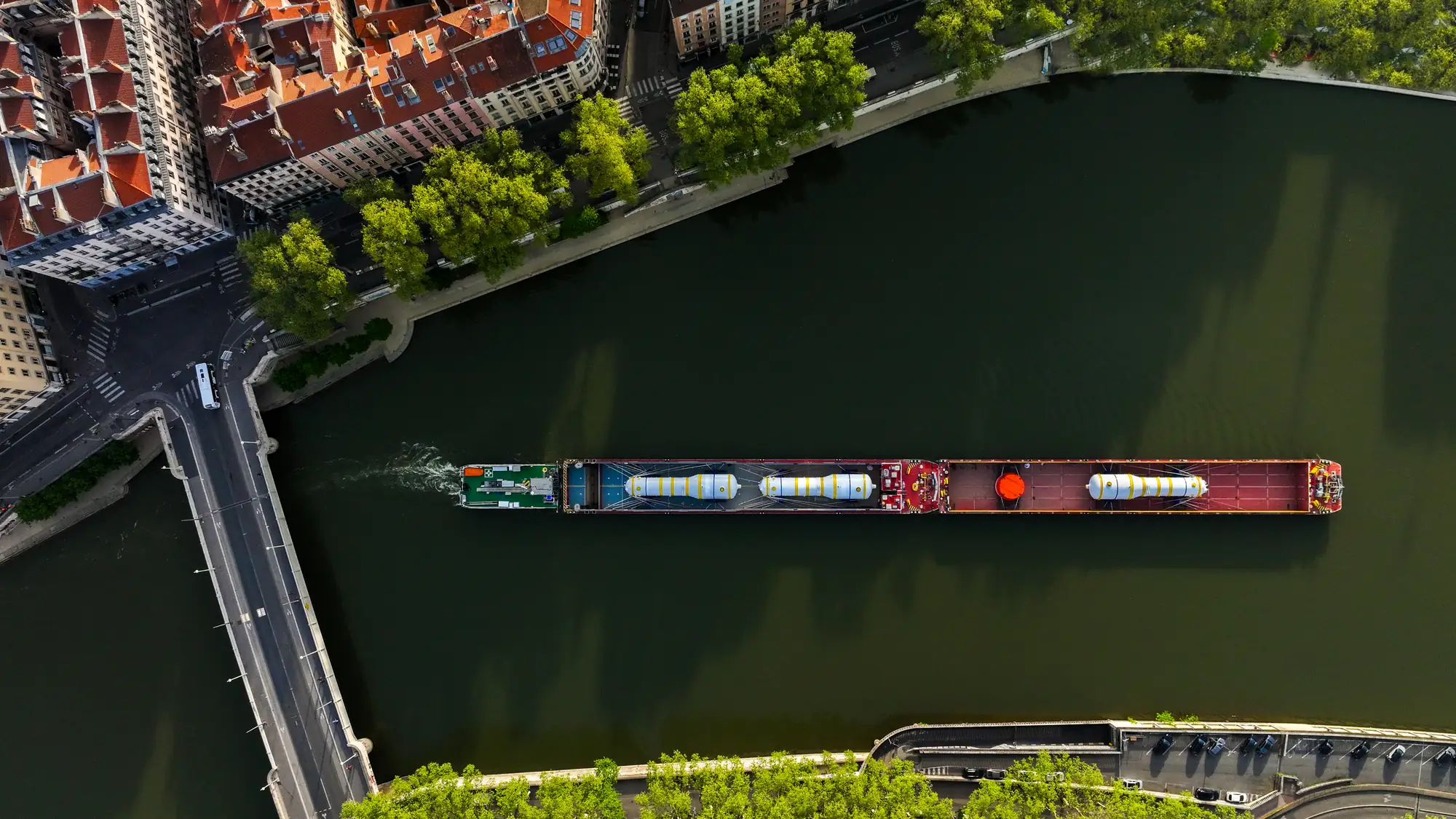 Vue aérienne à la verticale d’une barge fluviale transportant trois générateurs de vapeur, traversant une ville